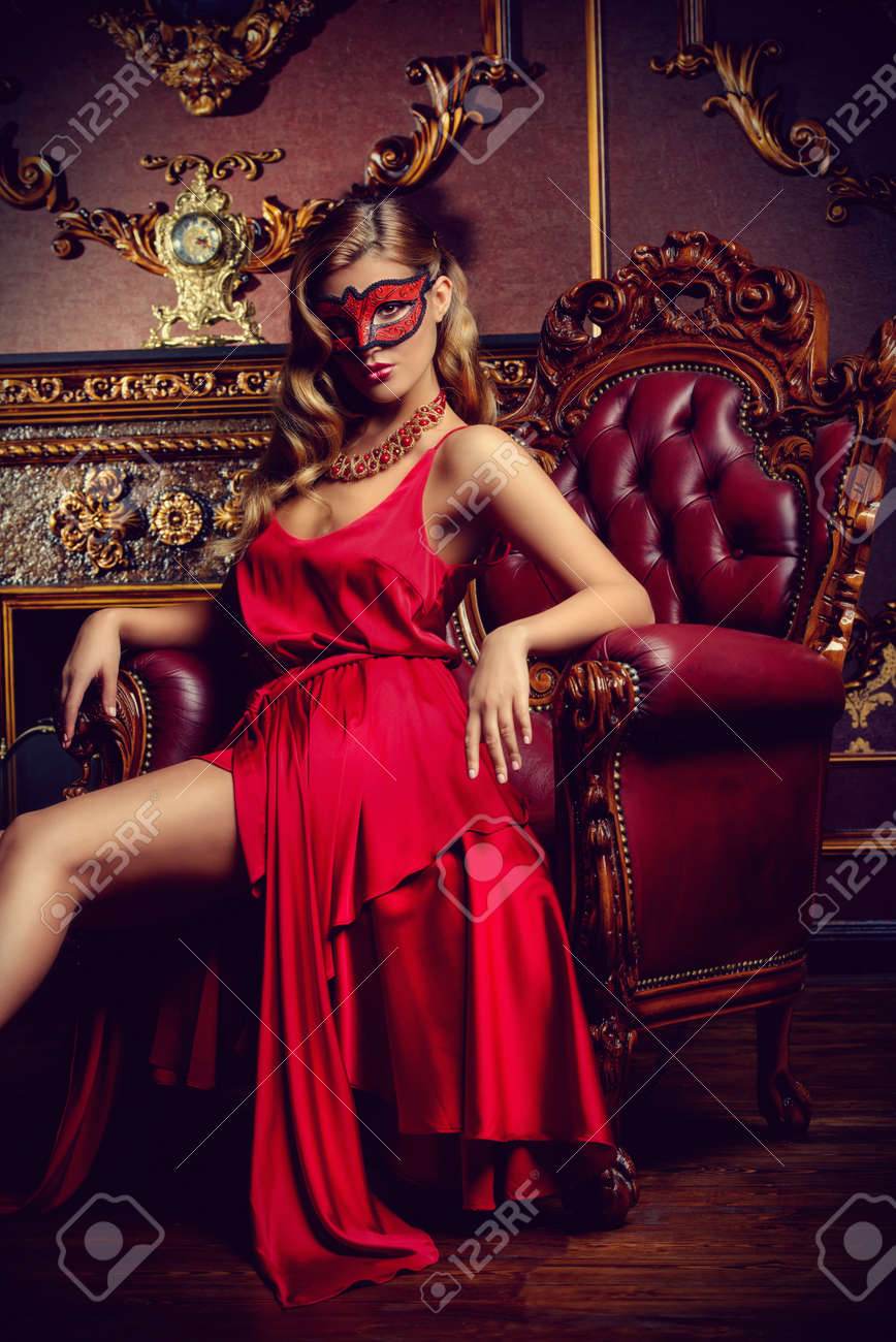 92295371-charming-elegant-woman-in-beautiful-red-dress-and-masquerade-mask-is-sitting-in-a-chair-in-a-luxury-.jpg